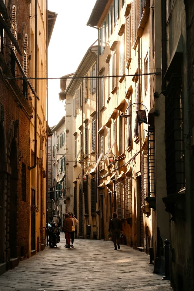 Travelers walking along a sunlit historic street in Lucca, Italy